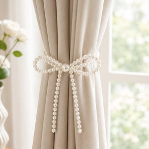May include: A beige curtain held back by a decorative tieback featuring a bow design made of white faux pearls. The centre of the bow is accented with a single, larger white pearl, and two long strands of pearls hang down from the centre knot.