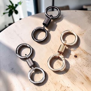 May include: Three metal keychains, each featuring two circular rings connected by a central cylindrical spring mechanism. One keychain is finished in a dark gunmetal gray, one in a polished silver tone, and one in a bright gold tone finish.