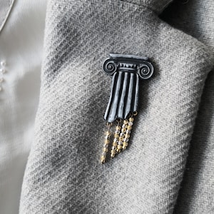 Può includere: Una spilla a forma di colonna architettonica grigio scuro con un capitello caratterizzato da volute a spirale. La base della colonna presenta quattro catenelle pendenti composte da piccole perle metalliche testurizzate dorate e argentate.