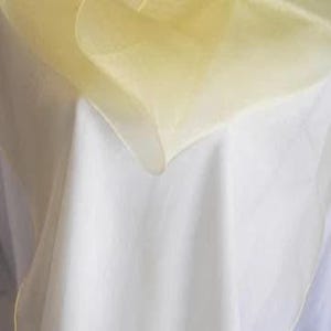 May include: A sheer, pale yellow fabric overlay draped over a solid white base cloth. The translucent, lightweight material creates soft folds and layered textures, showcasing a contrast between the warm yellow hue and the bright white fabric.