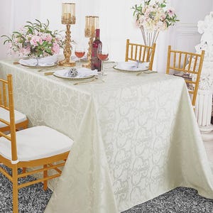 May include: A rectangular tablecloth in a light cream color featuring a tonal damask pattern. The table is set with white plates, gold-rimmed glassware, a wine bottle, two tall gold candle holders, and a floral centerpiece with pink flowers.