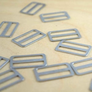 May include: A collection of silver-colored metal rectangular sliders, each featuring a central bar that divides the frame into two distinct openings. These hardware components are designed for adjusting straps on bags, backpacks, or various garments.