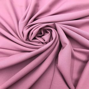 May include: A solid mauve-colored fabric featuring a smooth, matte texture. The material is arranged in a series of deep, flowing folds and gathers that create a dynamic, swirling pattern across the entire surface of the textile.
