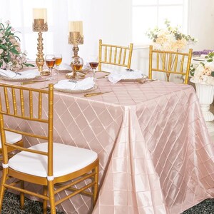 May include: A rectangular table covered with a blush pink satin tablecloth featuring a quilted diamond pattern. The table is set with white napkins, clear wine glasses, and gold candle holders. Gold chairs with white cushions surround the table.