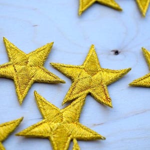 May include: A collection of five-pointed star patches featuring a vibrant gold thread embroidery with a dense, textured stitch pattern. Each star is individually crafted with a distinct sheen, showcasing a uniform metallic yellow color throughout.
