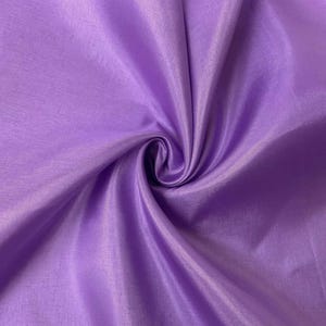 May include: A solid light purple fabric with a subtle sheen and a fine, woven texture. The material is gathered into a central swirl, creating soft, radiating folds and shadows that highlight the smooth, reflective surface of the textile.