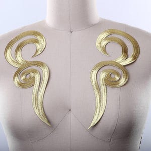 May include: A pair of symmetrical gold-colored embroidered appliques featuring swirling, elongated scroll patterns. These metallic-thread patches are positioned on a neutral-toned mannequin torso, showcasing a reflective, textured finish.