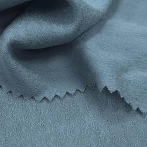 May include: A dusty blue fabric featuring a subtle, repeating geometric pattern with a textured, raised surface. The material is folded, creating soft drapes and shadows, with a zigzag-cut edge visible along the top layer of the textile.