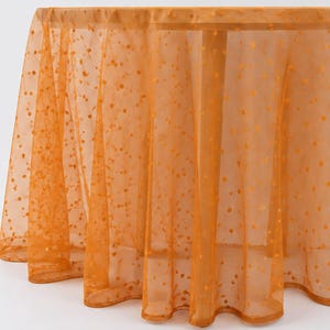 May include: A sheer orange tablecloth featuring a delicate dotted pattern throughout the fabric. The lightweight material drapes over a round surface, creating soft folds and ripples that highlight the translucent texture and vibrant color.