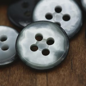 May include: A collection of round, four-hole buttons featuring a smooth, glossy finish with a marbled gray and black color pattern. Each button has a slightly raised outer rim and a recessed center area where the four circular holes are positioned.