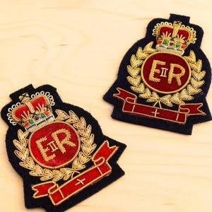 May include: Two embroidered patches featuring a dark blue felt base with a central red circle containing the gold embroidered letters ER II. Each patch displays a gold crown above the circle, gold laurel leaves, and a red ribbon banner at the base.