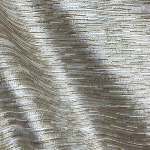 May include: A fabric with a textured surface featuring horizontal, raised white and beige threads woven into a repeating pattern. The material displays soft, undulating folds that create depth and highlight the interplay of light and shadow.