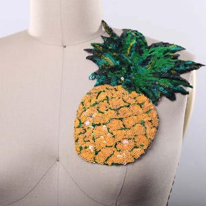 May include: A pineapple-shaped patch covered in sequins. The body of the pineapple is made of bright orange sequins, while the leafy top is crafted from dark green and black sequins, creating a textured and reflective surface on the fabric.