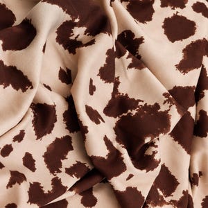 May include: A fabric featuring a repeating pattern of irregular dark brown spots against a light beige background. The material is draped to create soft folds and shadows, highlighting the texture and the contrast between the dark and light colors.