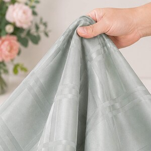 May include: A light sage green fabric featuring a subtle, tone-on-tone woven plaid pattern. The material has a smooth, slightly reflective finish and is draped in soft folds, showcasing the intricate grid design across the surface of the textile.
