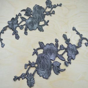 May include: Two dark gray floral embroidered patches featuring detailed stitching of blooming flowers, buds, and leafy stems. These decorative appliques are designed with a symmetrical branching pattern, showcasing intricate thread work and texture.