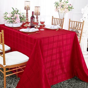 May include: A rectangular tablecloth in a deep red hue featuring a subtle, woven grid pattern. The fabric drapes over a table set with white plates, folded white napkins, gold-toned candle holders, and a wine bottle, surrounded by gold chairs.