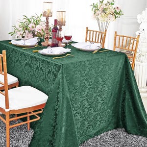 May include: A rectangular dark green tablecloth featuring a tonal damask pattern. The table is set with white napkins, gold-rimmed plates, gold-toned metal candle holders, wine glasses, and a wine bottle, surrounded by gold-toned wooden chairs.