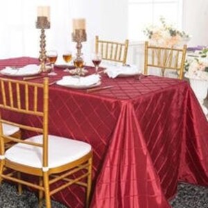 May include: A deep red rectangular tablecloth with a subtle diamond grid pattern draped over a table. The table is set with white plates, clear wine glasses, and two tall ornate gold candle holders with cream-colored pillar candles.