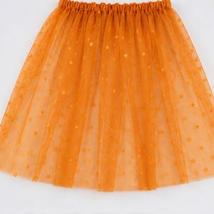 May include: An orange tulle skirt featuring a gathered elastic waistband and a sheer, layered fabric construction. The skirt is decorated with a repeating pattern of small, circular polka dots throughout the translucent, lightweight material.