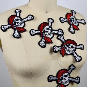 May include: Four embroidered patches featuring a white skull with a black eye patch and a red bandana, positioned over two crossed white bones. Each patch displays a detailed skull design with black outlines and shading on a white background.