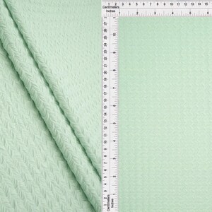 May include: A light mint green fabric featuring a textured herringbone pattern. A white ruler is positioned vertically along the center, showing measurements in both inches and centimeters, with the scale extending up to 27 centimeters or 10.5 inches.