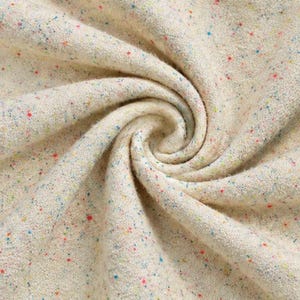 May include: A cream-colored fabric with a textured, fuzzy surface featuring a scattered confetti pattern of tiny red, blue, yellow, and green specks. The material is arranged in a swirling, spiral fold that creates deep shadows and highlights.