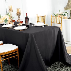 May include: A rectangular black tablecloth draped over a dining table set for a formal event. The table features white plates, gold-toned metal candle holders, a wine bottle, and gold-toned chairs with white cushions arranged around the table.