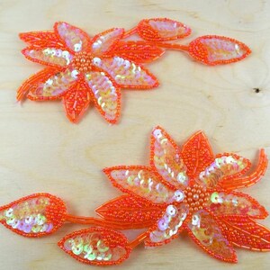 May include: Two orange floral appliques featuring intricate beadwork and iridescent sequins. Each flower has a central cluster of beads, radiating petals filled with orange seed beads and shimmering sequins, and two attached stems with leaf-shaped buds.