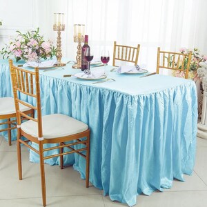 May include: A rectangular table covered with a light blue, floor-length pleated tablecloth, surrounded by gold-colored chairs with white cushions. The table is set with wine glasses, plates, silverware, and two tall gold candle holders.