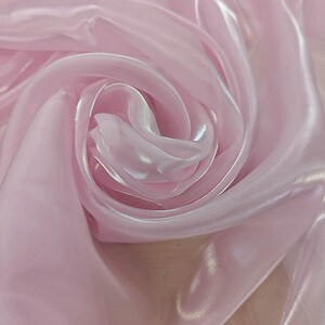 May include: A light pink, semi-sheer fabric with a smooth, lustrous finish. The material is arranged in soft, flowing folds that create a swirling, circular pattern, highlighting the reflective quality and delicate texture of the textile surface.