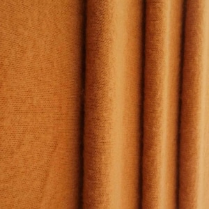 May include: A close-up view of a solid, warm-toned orange fabric featuring a soft, fine-knit texture. The material is arranged in vertical folds, creating subtle shadows and highlights that emphasize the consistent color and knit pattern throughout.