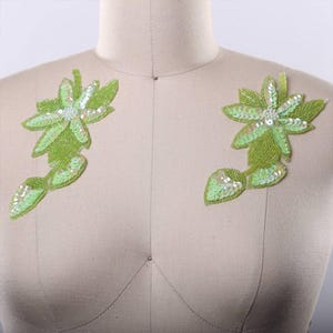 May include: Two matching lime green floral appliques featuring intricate beadwork and iridescent sequins. Each piece displays a central flower shape with pointed petals and attached leaves, crafted with light green beads and shimmering accents.