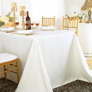 May include: A rectangular table covered with a solid white tablecloth, set with white plates, clear wine glasses, and gold-toned metal candle holders. Two gold-toned chairs with vertical slats are positioned at the table, and a wine bottle is present.