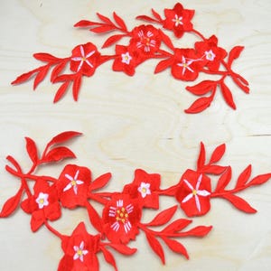 May include: Two red floral embroidered patches featuring detailed stitching of blossoms and leaves. Each patch displays multiple five-petaled flowers in vibrant red thread with white and yellow centers, connected by stems and pointed foliage.