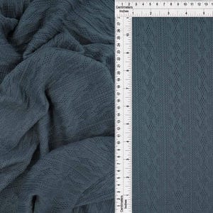 May include: A dark blue fabric featuring a vertical cable knit pattern with diamond-shaped textures. A white ruler is positioned along the side, showing the fabric width spans approximately 8 inches or 20 centimeters across the flat section.