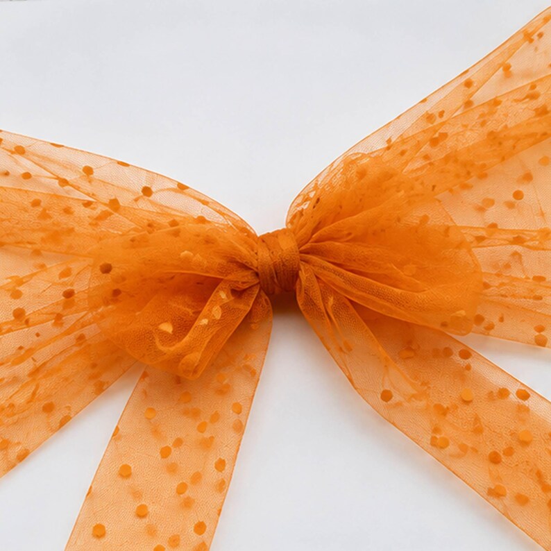 May include: A bright orange hair bow crafted from sheer, translucent tulle fabric. The material features a repeating pattern of small, raised, circular dots throughout the entire surface. The bow is tied in a classic shape with two long, flowing tails.