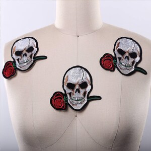 May include: Three embroidered patches featuring a white human skull with dark eye sockets and a red rose held between the teeth. Each patch has a black border and detailed stitching, showcasing a skull with a rose stem extending to the side.