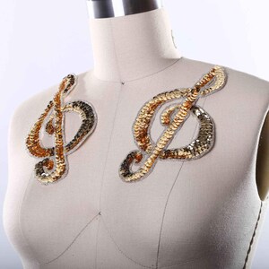 May include: Two musical treble clef appliques featuring gold and silver sequins. Each decorative patch is shaped like a G-clef, with a metallic finish and a white stitched border, displayed on a neutral-colored mannequin torso for clear visibility.