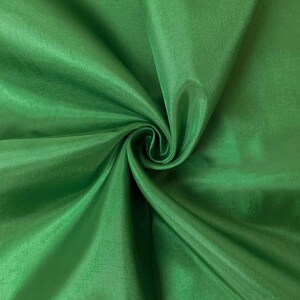 May include: A vibrant emerald green fabric with a smooth, slightly lustrous finish. The material is gathered in a central swirl, creating deep, flowing folds and shadows that highlight the consistent color and fine texture of the textile surface.