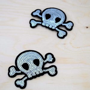 May include: Two skull and crossbones patches featuring silver sequins arranged on a black felt backing. Each patch displays a detailed skull design with hollow eye sockets and a jawline, flanked by two crossed bones, creating a high-contrast look.
