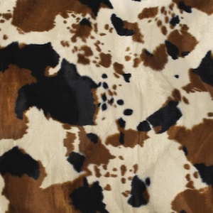 May include: A soft fabric featuring a tri-color cow print pattern with irregular patches of black, dark brown, and cream white. The surface has a short, plush texture that creates subtle shadows and highlights across the organic, spotted design.
