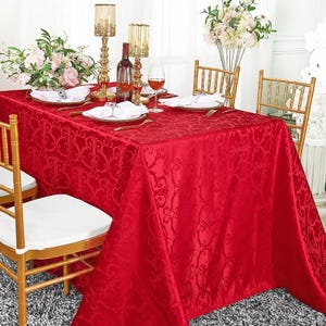 May include: A rectangular red tablecloth with a tonal damask pattern covering a dining table. The table is set with white plates, white napkins, gold-toned candle holders, a wine bottle, wine glasses, and floral arrangements, surrounded by gold chairs.