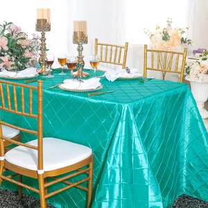 May include: A rectangular table covered with a vibrant teal satin tablecloth featuring a stitched diamond grid pattern. The table is set with white plates, folded white napkins, gold-toned candle holders, and clear glassware, surrounded by gold chairs.