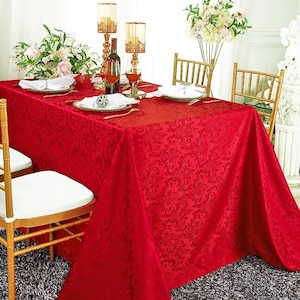 May include: A rectangular red tablecloth featuring a tonal damask pattern draped over a dining table. The table is set with white plates, gold-rimmed chargers, gold candle holders, a wine bottle, and floral arrangements, surrounded by gold chairs.