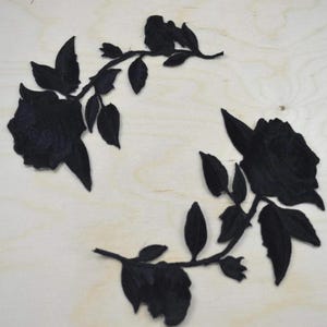 May include: Two black embroidered rose patches featuring detailed floral blooms, stems, and leaves. Each patch displays a single rose head connected to a curved stem with multiple pointed leaves, rendered in solid black thread embroidery.