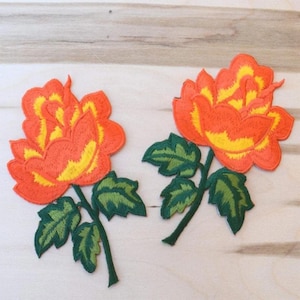 May include: Two embroidered fabric patches, each featuring a vibrant orange rose with yellow inner petals and dark green leaves on a stem. The rose designs are detailed with dense stitching, showcasing a layered petal structure and serrated foliage.
