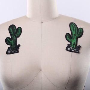 May include: Two identical cactus-shaped patches featuring green sequins and black embroidered outlines. Each patch depicts a saguaro-style cactus standing on a small, textured black base with white accents, attached to a beige fabric mannequin torso.