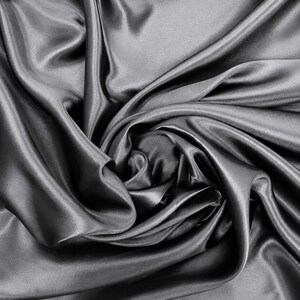 May include: A dark gray fabric with a smooth, lustrous surface and a high-sheen finish. The material is draped in deep, flowing folds that create a series of light and shadow patterns across the surface, highlighting the soft, fluid texture.