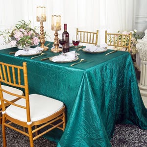 May include: A rectangular emerald green crinkled tablecloth draped over a table set with white napkins, wine glasses, a wine bottle, and two gold candle holders. Four gold chairs with white cushions are positioned around the table.